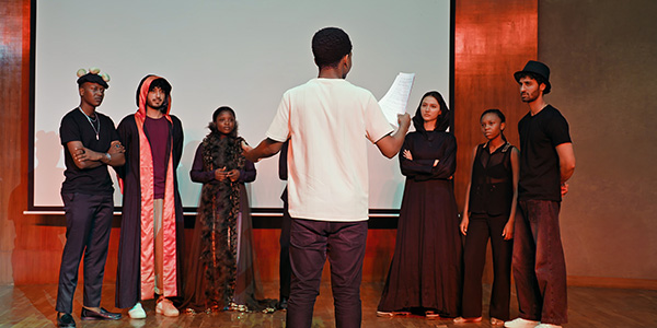 A group of students in various costumes standing on a stage during a rehearsal.